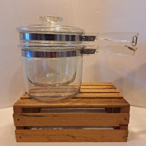 May include: A clear glass double boiler with a lid and two clear handles. The glass pot has a silver metal band around the top and is sitting on a small wooden pallet.