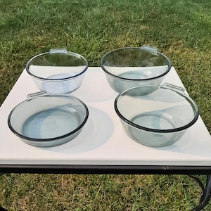 May include: Four clear glass baking dishes with handles on a white table. The dishes are all different sizes and have a slight blue tint.