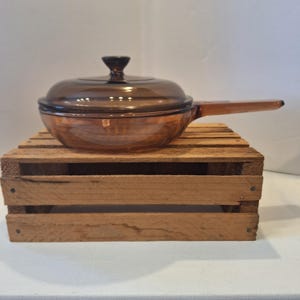 May include: A vintage amber-colored glass saucepan with a matching lid and a long handle. The cookware is displayed on a rustic wooden crate, showcasing its classic design. This kitchenware is perfect for cooking.