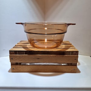 May include: A round, amber-colored glass casserole dish with two small, integrated side handles. The bowl features a smooth, translucent finish and a distinct horizontal ridge encircling the upper portion of the body, providing a simple, functional design for kitchen use.