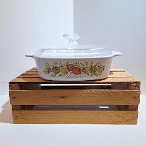 May include: White casserole dish with a clear glass lid. The dish features a colorful fruit and vegetable design with the text "L'échalote" below the design. The dish is sitting on a wooden crate.