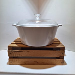 May include: A white casserole dish with a clear glass lid. The dish is sitting on a small wooden crate. The casserole dish has two small handles on either side. The lid has a small knob on top.