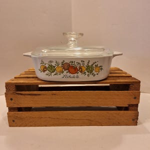 May include: White casserole dish with a glass lid. The dish has a floral design with lemons, tomatoes, and herbs. The text "L'Echalote" is printed on the dish.