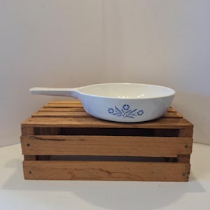 May include: White ceramic skillet with a long handle and blue floral pattern. The skillet is sitting on a wooden crate. The floral design is a simple, stylized arrangement of flowers and leaves.