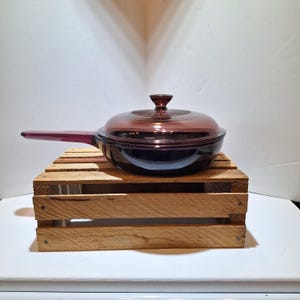May include: A vintage, dark purple glass skillet with a matching lid. The skillet has a long handle and sits on a wooden crate. The lid has a knob for easy handling. The cookware is designed for stovetop use.