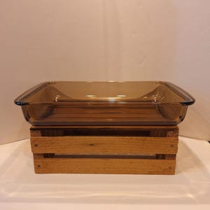 May include: A rectangular, amber-colored glass baking dish with rounded corners. The dish is sitting on a wooden crate. The glass has a slight tint, and the overall design is simple and functional. Ideal for baking and serving.