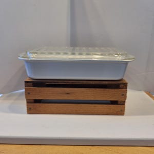 May include: A rectangular, light blue casserole dish with a clear glass lid. The dish is resting on a small, wooden crate. The crate has a natural wood finish and is made of horizontal slats.