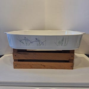 May include: A rectangular, white baking dish with a floral design in blue and green. The dish has handles on each side and is sitting on a wooden crate. The floral design features stylized irises.