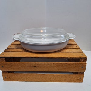 May include: White ceramic baking dish with a glass lid. The dish has two handles on the sides and is sitting on a wooden crate.