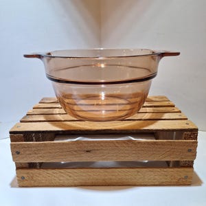 May include: A translucent amber-colored glass bowl featuring a wide, circular rim and two integrated side handles. The bowl has a smooth, rounded base and a distinct horizontal ridge encircling the upper section of the glass vessel.