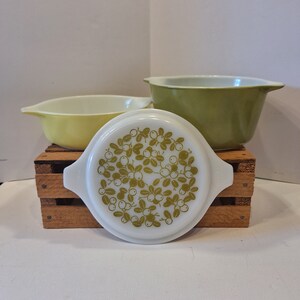May include: Two white casserole dishes with green floral designs and a matching lid. The dishes are on a wooden crate.