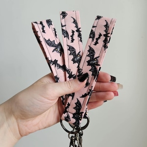 May include: Three pink fabric keychains with black bat silhouettes. The keychains have silver key rings.