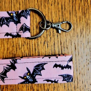 May include: Pink fabric key chain with a black metal ring and clasp. The fabric has a repeating pattern of black bats with yellow eyes on a pink background.