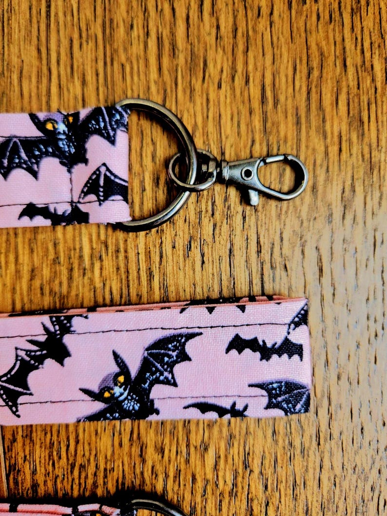 May include: Pink fabric key chain with a black metal ring and clasp. The fabric has a repeating pattern of black bats with yellow eyes on a pink background.