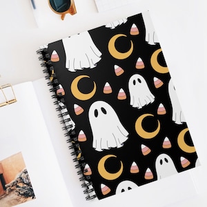 Może przedstawiać: Czarny notes w spirali z motywem Halloween. Okładka przedstawia powtarzający się wzór białych duchów, żółtych półksiężyców i pomarańczowo-różowego kukurydzy.