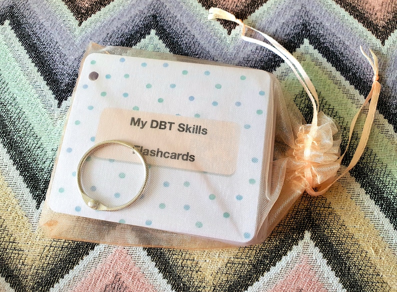 May include: A set of white DBT Skills flashcards with blue polka dots, packaged in a peach-colored organza bag with a silver ring binder. The text on the flashcards reads "My DBT Skills Flashcards".
