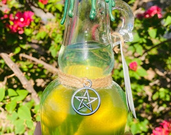 May include: A clear glass bottle filled with a yellow-green liquid, sealed with a cork and green wax. A silver pentagram charm hangs from a twine around the bottle's neck. The bottle has a handle and a tag.