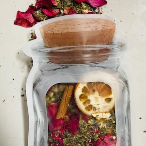 May include: A clear glass jar filled with dried herbs, rose petals, a cinnamon stick, and a dried lemon slice. The jar is sealed with a cork stopper and a clear plastic pouch. A pile of rose petals and herbs sits on top.