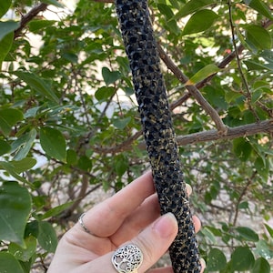 May include: A black candle with a textured surface, held against a backdrop of green leaves. The candle has a cylindrical shape and is adorned with small, dark embellishments and flecks of gold.