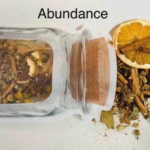 May include: A clear glass jar with a cork stopper, filled with dried herbs and spices. A slice of dried orange and cinnamon sticks are next to the jar. The word "Abundance" is written above the jar.