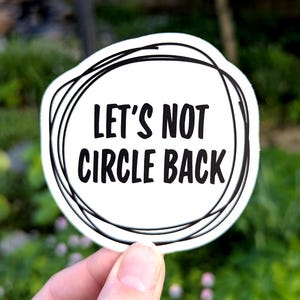 May include: A white sticker with the text "LET'S NOT CIRCLE BACK" in black, surrounded by a black scribble design. The sticker is held up against a blurred green background.