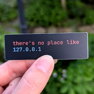May include: A black rectangular sticker with the text "there's no place like 127.0.0.1" in red and blue font. The sticker is held in a person's hand against a blurred green background.