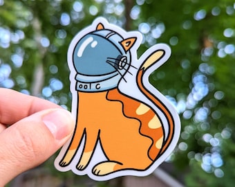 Puede incluir: Una pegatina de un gato atigrado naranja con un casco de astronauta azul con una visera blanca. El gato está sentado con la cola enroscada alrededor de su cuerpo. La pegatina está sobre un fondo blanco.