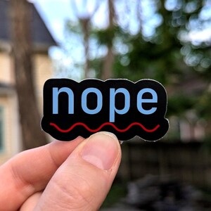 Puede incluir: Una pegatina negra con la palabra "nope" en letras azul claro. Una l&iacute;nea ondulada roja est&aacute; debajo de la palabra. La pegatina se sostiene en una mano sobre un fondo borroso al aire libre.