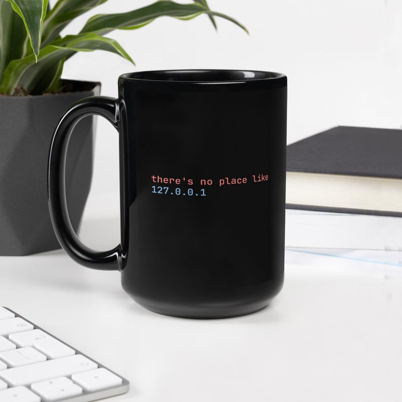 May include: Black coffee mug with the text "there's no place like 127.0.0.1" in red and blue. The mug has a large handle and is on a white surface. A plant and a book are in the background.