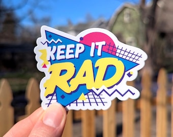 May include: A colorful sticker with a retro 80s design. It features the text "Keep it Rad" in a bold, yellow font with blue and pink accents. The sticker has a geometric pattern with zigzags and grid lines.