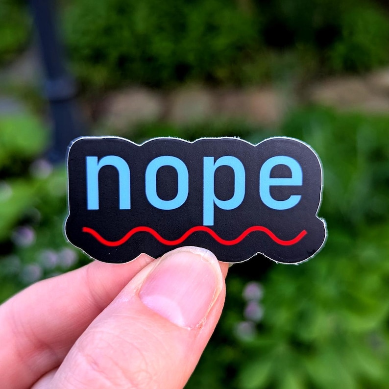 Puede incluir: Una pegatina negra con la palabra "nope" en letras azul claro. Una l&iacute;nea ondulada roja est&aacute; debajo de la palabra. La pegatina se sostiene en una mano sobre un fondo verde borroso.