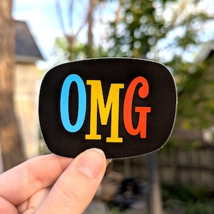 May include: A black sticker with the letters "OMG" in blue, yellow, and red. The sticker has rounded corners and is held up by a hand against a blurred background of green foliage.