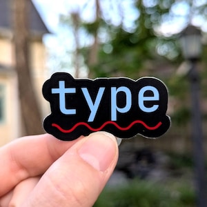 Könnte beinhalten: Ein schwarzer Aufkleber mit dem Wort "type" in hellblauen Buchstaben. Am unteren Rand des Aufklebers befindet sich eine rote Wellenlinie. Der Aufkleber wird zwischen zwei Fingern gehalten.