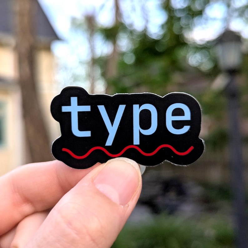 May include: A black sticker with the word "type" in light blue letters. A red wavy line is at the bottom of the sticker. The sticker is held between two fingers.