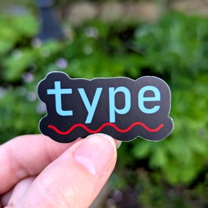 May include: A black sticker with the word "type" in light blue, and a red wavy line at the bottom. The sticker is held in a hand against a blurred green background.
