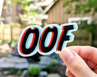 May include: A sticker with the word "OOF" in a 3D, red, blue, and black design. The sticker is on a hand.