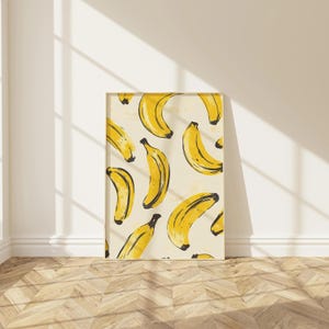 Op de afbeelding: Een ingelijste print met een herhalend patroon van gele bananen op een crèmekleurige achtergrond. De bananen hebben zwarte steeltjes en zijn in een verspreide, artistieke stijl gerangschikt. De print wordt gepresenteerd tegen een witte muur en een houten vloer.