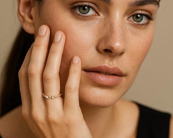 May include: A gold-colored open ring with two small, clear gemstones. The ring has a textured, rope-like design. The ring is worn on a finger. The background is a neutral beige color.