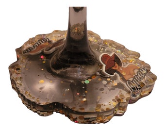 May include: A clear glass wine glass with a gold glitter base. The base has a sticker with a woman wearing a red hat and the word "Queen" written on it.