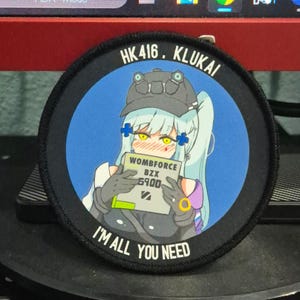May include: A circular patch with a black border featuring an anime-style illustration. The image shows a character with light blue hair, yellow eyes, and a military-style cap. Text includes "HK416. KLUKAI", "WOMBFORCE BZX 6900", and "I'M ALL YOU NEED".