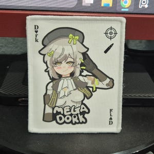 May include: White rectangular patch with a cartoon illustration of a character wearing a hat and holding a gun. The text "MEGA DORK" is below the character. The patch has the letters "DORK" and "KLAD" on the sides.
