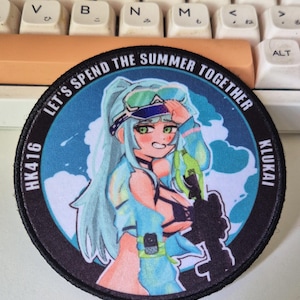 May include: A round patch featuring an anime-style character with blue hair and a water gun, set against a blue sky background. Text around the edge reads "LET'S SPEND THE SUMMER TOGETHER" and "KLUKAI". The patch also has the text "HK416".