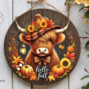 Può includere: Un cartello rotondo in legno con una mucca delle Highlands che indossa un cappello a quadri decorato con girasoli. Il cartello è decorato con zucche, foglie d'autunno e le parole "Hello Fall". Il cartello misura 30,48 x 30,48 cm.