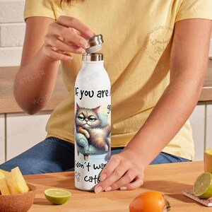May include: White water bottle with a screw-on lid. The bottle features a grumpy cat illustration and the text "If you are gonna be grumpy, don't want cats". The bottle is on a wooden surface.
