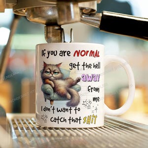 May include: White ceramic mug with a humorous cat illustration and text. The cat is lounging on a small armchair. The mug has the text "If you are NORMAL get the hell away from me I don't want to catch that SHIT."