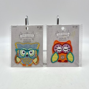 May include: Two iron-on applique patches featuring colorful owls. The owl on the left is wearing glasses and has a blue, green, and orange color scheme. The owl on the right has a pink, orange, and yellow color scheme. Both patches are packaged on white cards with the text "Sew-ology Iron-on Applique" and a price of $1.99.
