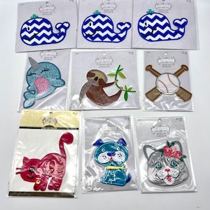 May include: A collection of iron-on patches featuring various designs, including a blue whale, a sloth, a baseball and bats, a narwhal, a cat, a dog, and a cat with a flower crown. The patches are packaged individually on white cards with the brand name "Sew-ology" and the text "Iron-on Applique".