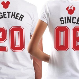 Op de afbeelding: Twee witte T-shirts met rode nummers en zwarte tekst. Het shirt aan de linkerkant zegt "TOGETHER 20" en het shirt aan de rechterkant zegt "SINCE 06". Beide shirts hebben een rood Mickey Mouse hoofd met zwarte contouren.