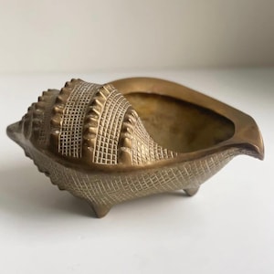 May include: A detailed, golden-toned seashell-shaped decorative bowl. The bowl features a textured surface with intricate patterns and rests on three small feet. The interior is smooth and open, perfect for small items.