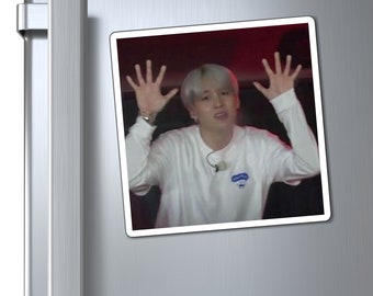 May include: A man with grey hair is making a surprised expression with his hands raised in the air. He is wearing a white t-shirt with a blue logo.